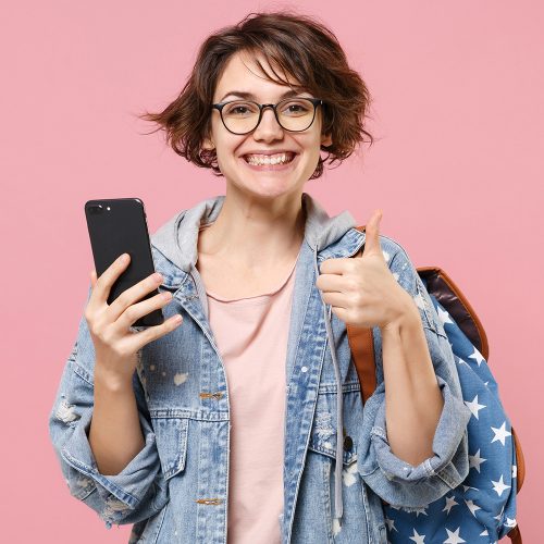Smiling woman student in denim clothes glasses backpack isolated on pastel pink background. Education in high school university college concept. Using mobile phone typing sms message showing thumb up